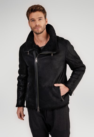 Uomo che indossa una giacca in shearling nera con colletto alto, chiusura a zip, tasche con zip e pantaloni neri casual su uno sfondo semplice.
