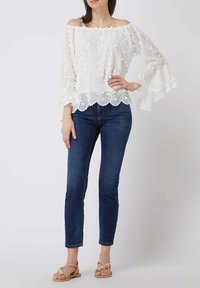 Blouse blanche à col bateau avec broderie florale et manches cloche volantées, associée à un jean bleu slim et des sandales marron clair.