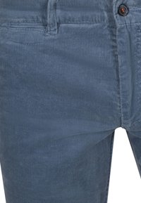 Close-up van een blauwe corduroy broek met zicht op de voorzak, tailleband met riemlus en een donkere knoop met oranje stiksel.