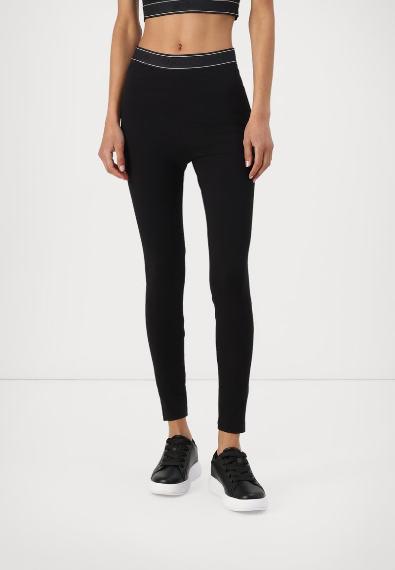 Fekete leggings magas derékkal, kontrasztos logósárral. Fekete cipővel, fehér talppal párosítva. Simaságával és rugalmas anyagával kiemelkedő.