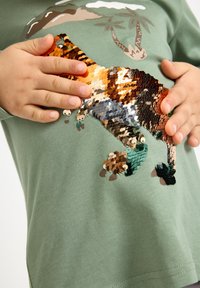 Grøn bomulds langærmet skjorte med et farverigt paliet-dinosaur-design på fronten. Glat tekstur med afslappet pasform.