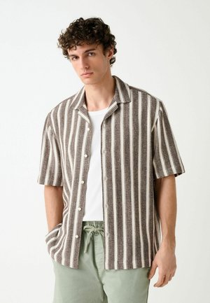 Junger Mann mit lockigem Haar, der ein kurzärmeliges, braun-weiß vertikal gestreiftes Hemd über einem weißen Oberteil und hellgrünen Hosen trägt.