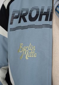 Veste bleu clair avec texte "PROH" en noir et "Berlin Mitte" brodé en or, portée sur un haut en tricot beige et une cravate rayée.