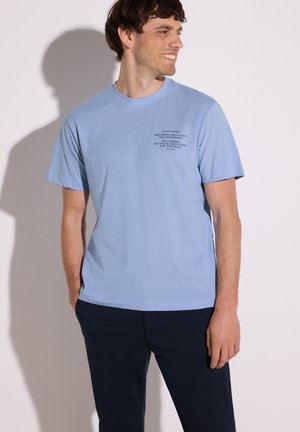 Homme souriant, portant un t-shirt bleu clair à manches courtes avec un texte noir sur la poitrine gauche, et un pantalon bleu foncé, mains dans les poches, fond clair.
