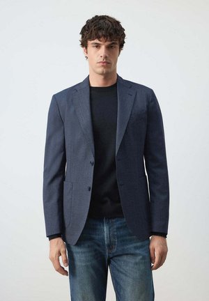 Hombre con una chaqueta azul oscuro sobre una camisa negra combinada con vaqueros azules, de pie frente a un fondo claro y liso.