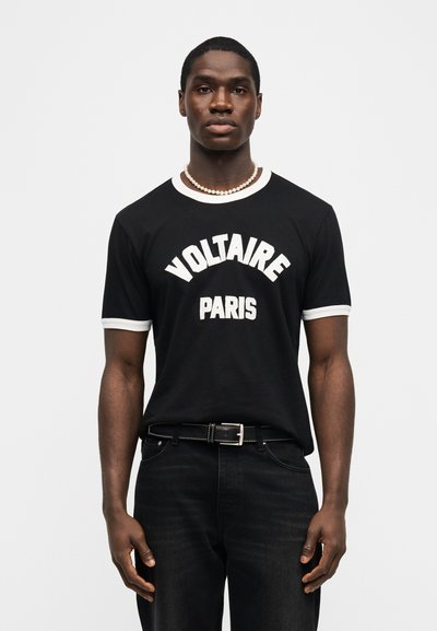 Μαύρος άνδρας που φοράει μαύρο μπλουζάκι με λευκή επιγραφή "VOLTAIRE PARIS", μαύρο τζιν, μαύρη ζώνη και μαργαριταρένιο κολιέ, στέκεται μπροστά σε απλό λευκό φόντο.