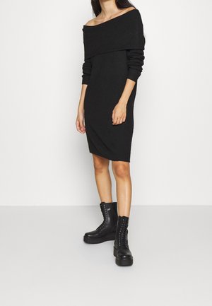 Kvinde iført en sort off-shoulder strikkjole kombineret med sorte snørebånd-støvler, stående mod en hvid baggrund.