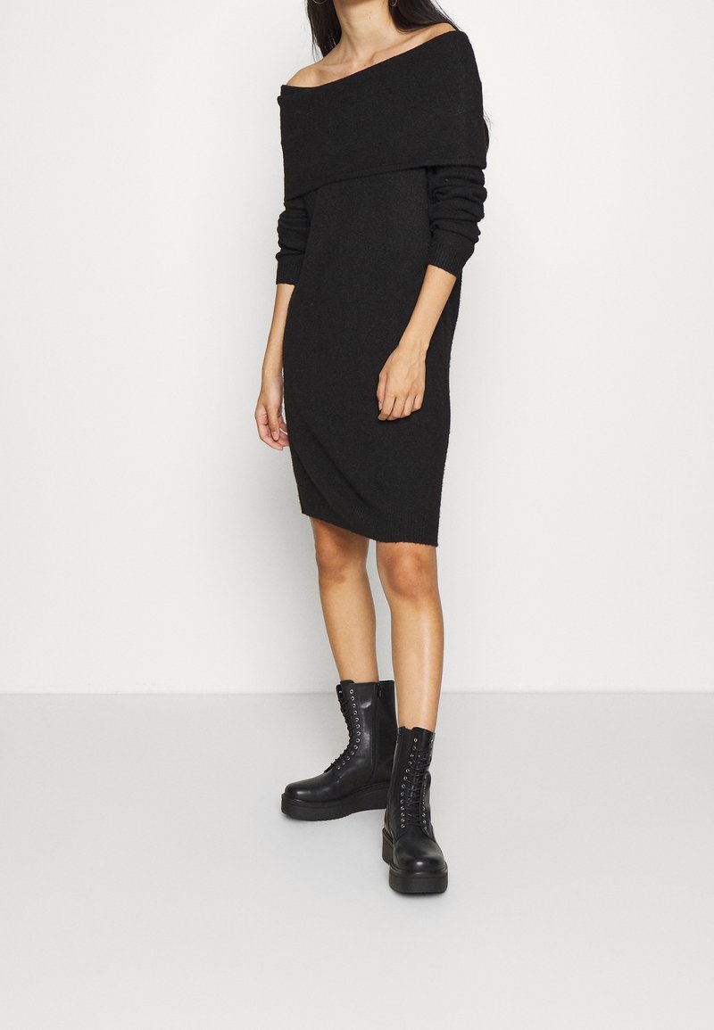 Femme portant une robe en maille noire avec épaules dénudées assortie à des bottines noires à lacets, debout devant un fond blanc.