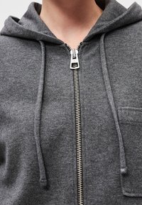 Gros plan d'une personne portant un sweat à capuche gris à fermeture éclair argentée avec cordons, montrant le cou et la partie supérieure de la poitrine.