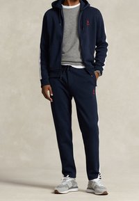 Marinträningsoverall med vita sidostänger, zip-up huvtröja och grå sweatshirt under. Texturerad tyg, byxor med dragsko och grå sneakers.