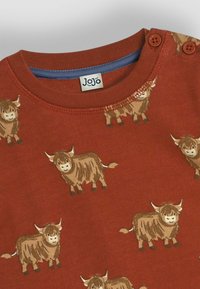 Roter Baumwollpullover mit einem wiederholten Muster von cartoonhaften Highland-Rindern, mit zwei Knöpfen an der Schulter und einem kontrastierenden blauen Kragen innen.