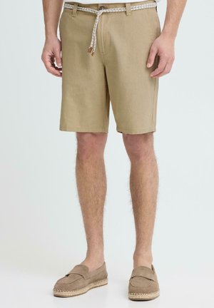 Man draagt beige knielange korte broek met een witte gevlochten riem en lichtbruine instapschoenen tegen een effen achtergrond.