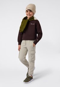 Smilende barn iført beige lue, mørk sweatshirt med grønn skjerf over skulderen, beige cargo bukser og olivengrønne sneakers som står foran en hvit bakgrunn.
