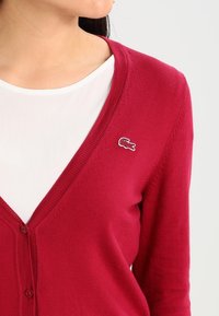 Crimson cardigan with a V-neck, button front, and Lacoste logo detail on the left side. Soft, knitted texture and fitted design.