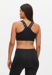 Musta urheilutoppi ristikkäisillä olkaimilla, joissa on logo-yksityiskohtia. Sileä, joustava kangas, joka on suunniteltu mukavuuden ja tuen tarjoamiseksi.