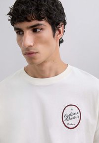 Camiseta de algodón blanca con un logo redondo en rojo oscuro y el texto "Scalpers Librarie". Cuello acanalado y textura suave.