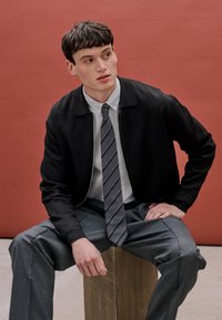 Veste zippée noire sur une chemise à rayures avec un col boutonné et une cravate grise. Pantalon gris avec un design texturé. Fond de couleur unie.