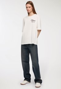 Biała, oversize'owa koszulka z krótkim rękawem i frontowym nadrukiem. Połączona z ciemnoniebieskimi, szerokimi dżinsami i białymi trampkami.