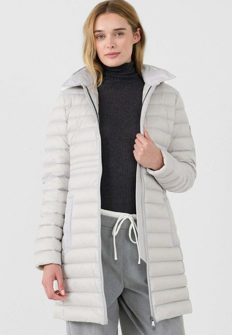 Lichtgrijze puffy jas met een rits aan de voorkant, geribbeld kraag en horizontale stiksels. Gepaard met een donkere top en lichtgrijze broek.