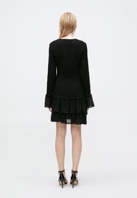 Vestido negro con mangas largas y acampanadas, cuerpo texturizado y falda de volantes en capas. El dobladillo presenta un detalle de encaje con forma de scalloped. Se lleva con tacones altos.