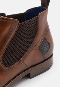 Bruine leren Chelsea-boot met gevlochten elastische panelen, een blauwe voering en een zwart logo-opdruk op de zijkant. Heeft een lage houten hak.