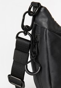 Detail einer schwarzen Schultertasche mit einem strukturierten Lederpanel, gewebtem Stoffgurt, Metallbeschlägen und einem Karabinerclip.