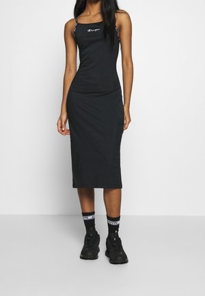 Kobieta ubrana w dopasowaną, czarną sukienkę midi Champion na cienkich ramiączkach, czarne trampki i czarne skarpetki z białym logo.