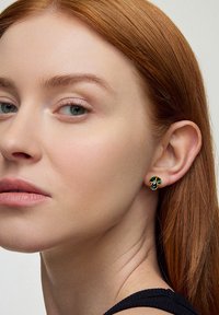 Złote kolczyki w kształcie liści z niebieskimi i zielonymi akcentami. Zbliżenie na ucho, ukazujące szczegółowy wzór i gładką teksturę na skórze.