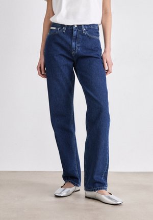 Persona vistiendo jeans azul oscuro de pierna recta, una camiseta blanca de manga corta y bailarinas plateadas, de pie sobre un suelo beige.