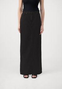 Kvinna iförd en svart ärmlös topp och en lång svart denimkjol, tillsammans med svarta öppna tåklackar, stående mot en vit bakgrund.