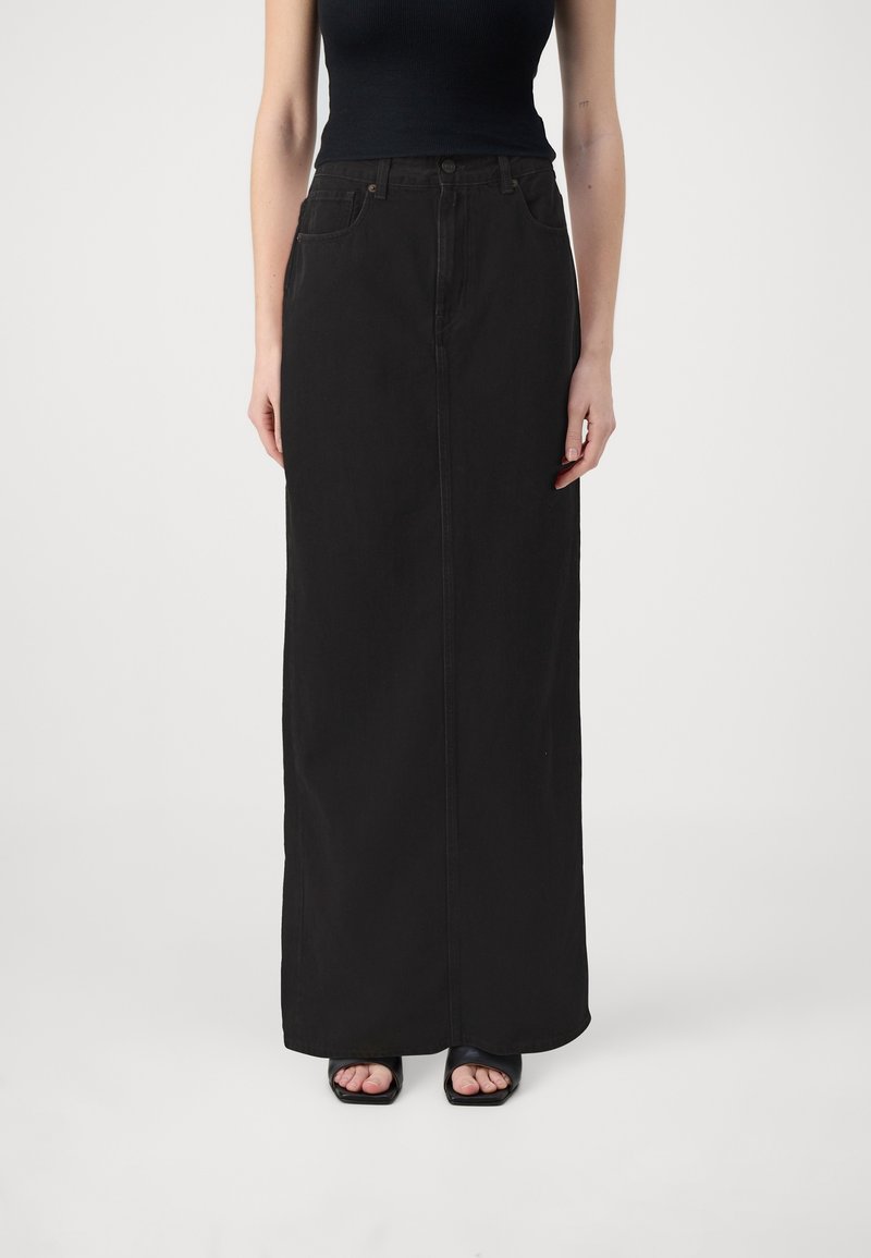 Kvinna iförd en svart ärmlös topp och en lång svart denimkjol, tillsammans med svarta öppna tåklackar, stående mot en vit bakgrund.