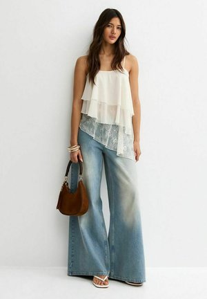 Vrouw draagt een witte gelaagde kanten top, wijde verwassen blauwe jeans, draagt een bruine handtas, met armbanden en witte sandalen.