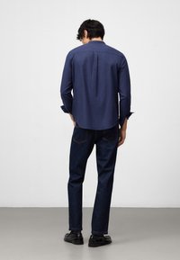 Maglietta a maniche lunghe blu navy con orlo dritto, dettaglio della piega posteriore, abbinata a jeans in denim scuro e scarpe nere, vista da dietro.
