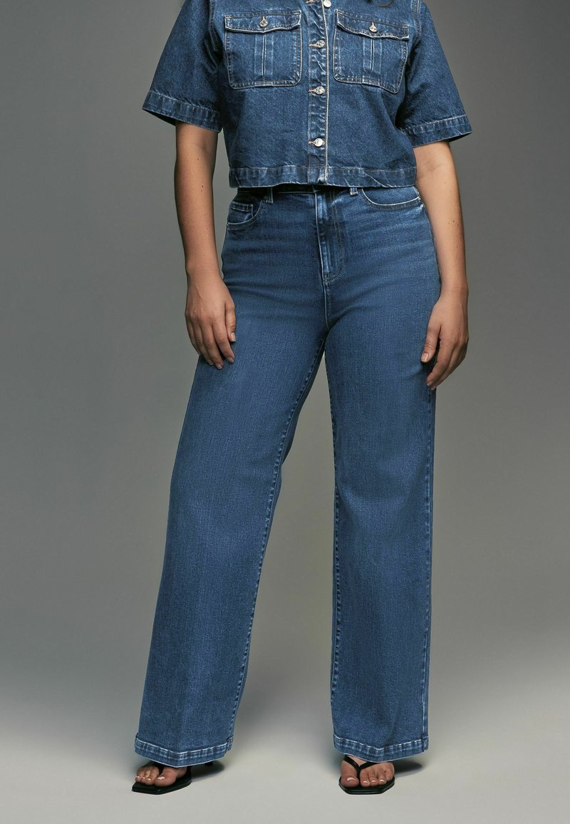 Veste en denim coupée avec deux poches poitrine et fermeture à boutons, associée à un jean en denim bleu taille haute et à jambes larges. Texture lisse.
