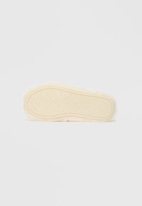 Crème-kleurige fuzzy pantoffelzool met een gewatteerde beige binnenzool met een herhaald logo patroon. Cirkelvormig en plat, lichtjes gewatteerd.