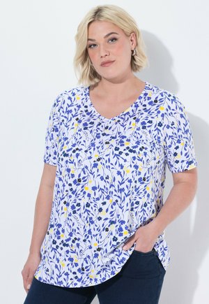 Lyshåret kvinde iført sølv hoops-øreringe og en hvid V-hals skjorte med blå og gule blomster, poserer med den ene hånd i lommen.