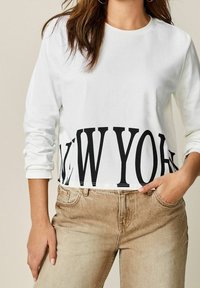 Frau trägt ein weißes Langarmshirt mit "NEW YORK" in großen schwarzen Buchstaben bedruckt und beigen hochtaillierten Jeans, die Hand in der Tasche.