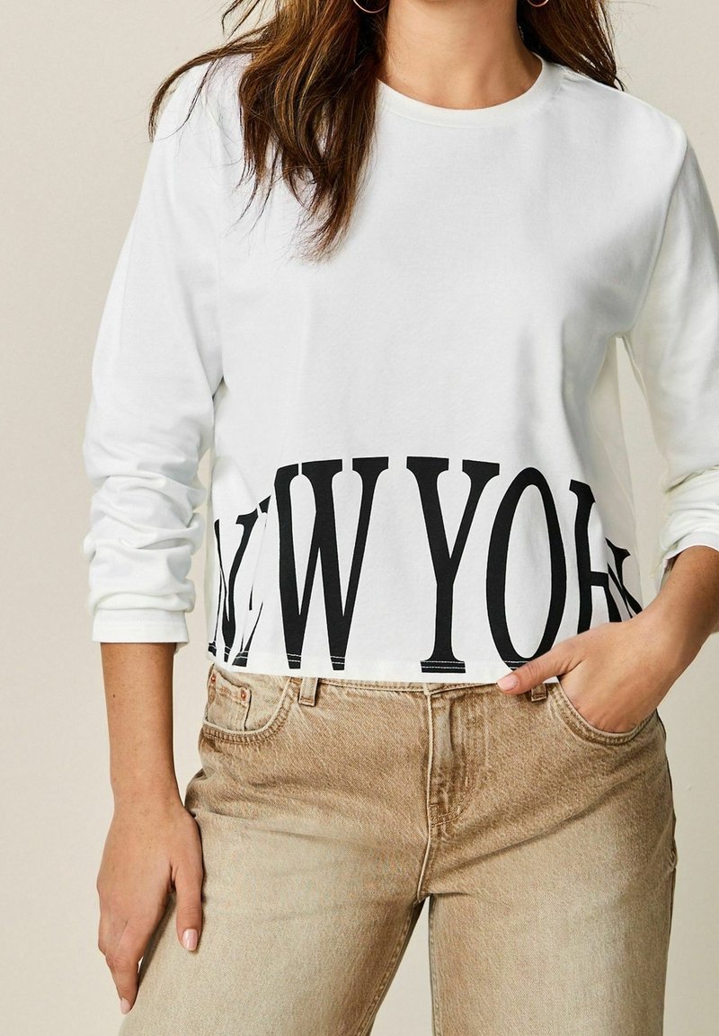 Frau trägt ein weißes Langarmshirt mit "NEW YORK" in großen schwarzen Buchstaben bedruckt und beigen hochtaillierten Jeans, die Hand in der Tasche.
