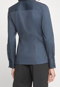 Camicia fitted blu navy con una texture liscia, maniche lunghe, cuciture sulle spalle e chiusura con bottone ai polsini. Design minimalista, senza motivi.