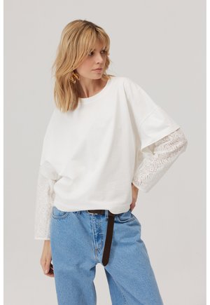 Kobieta w białym topie z długim rękawem z koronkowymi rękawami, niebieskich dżinsach z czarnym paskiem i złotych kolczykach w kształcie liści, patrząca w bok.