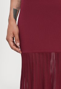 Abito bordeaux con un corpetto fitted e un orlo frangiato realizzato in materiale trasparente, con una superficie lavorata a maglia liscia sopra.