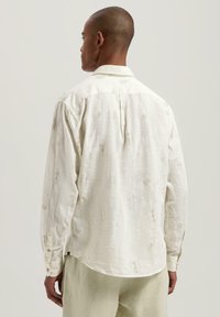 Hvit langermet skjorte laget av teksturert stoff med subtile bladmønstre, knappede mansjetter og buet falm. Synlig bakfolddetalj.