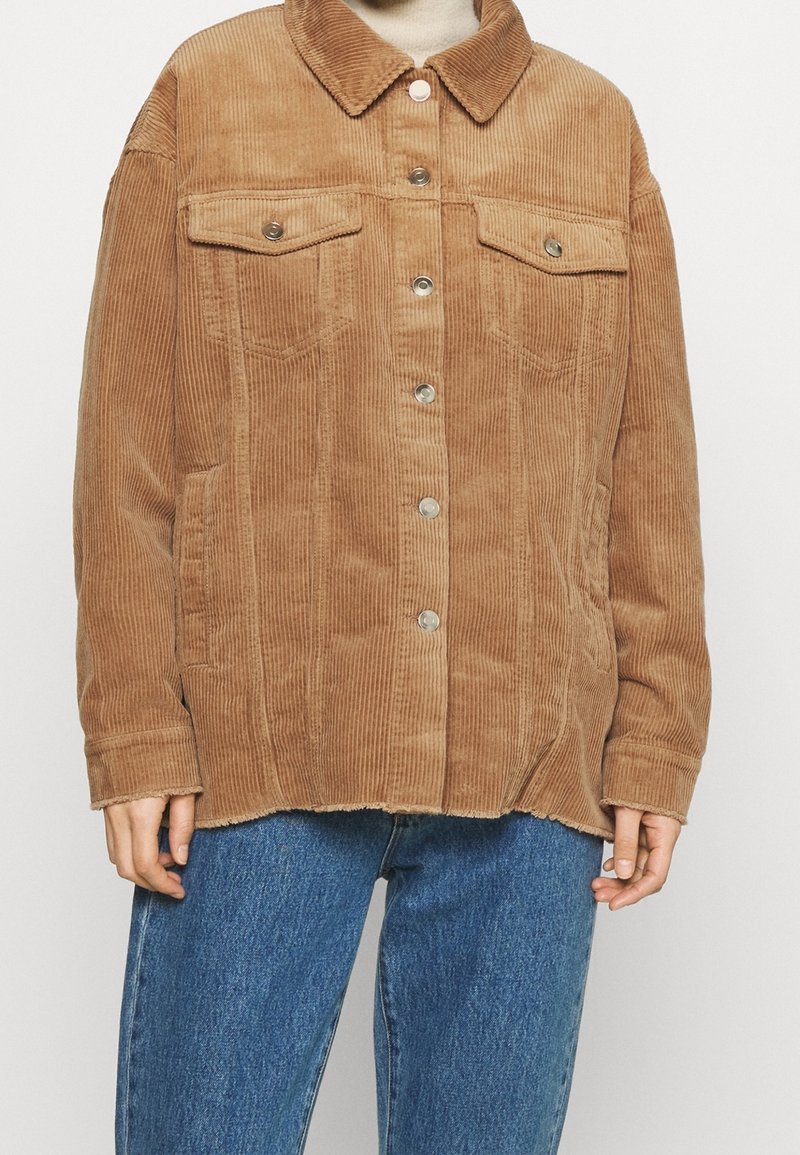 Veste en velours côtelé marron clair à boutons, avec col et poches poitrine à rabat, portée sur un col roulé crème et associée à un jean bleu.
