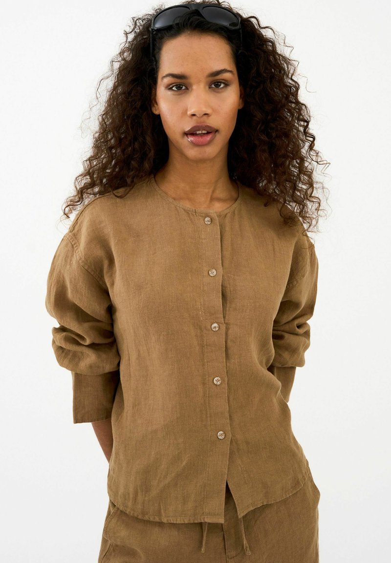 Jeune femme aux cheveux bouclés portant une chemise marron à boutons et un pantalon assorti, lunettes de soleil reposant sur sa tête, fond neutre.