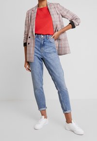 Blazer à carreaux rouge, noir et blanc avec fermeture à deux boutons. T-shirt rouge et jeans taille haute bleu clair, associés à des baskets blanches.