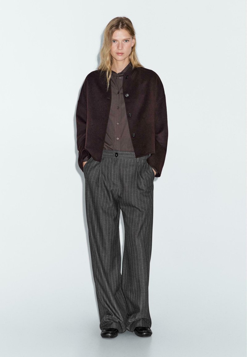 Dark brown cropped jacket over a dark shirt, paired with gray pinstriped wide-leg trousers and black shoes, against a neutral background.