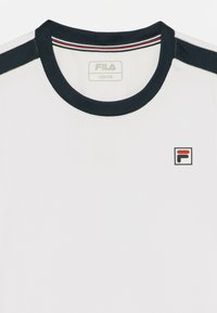 T-shirt blanc avec un col rond bleu marine. Présente des accents d'épaule bleu marine et un petit logo Fila rouge et noir sur la poitrine. Texture lisse.
