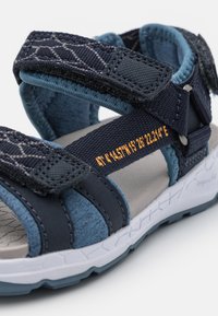 Sandali per bambini blu navy e grigi con chiusure in Velcro, tessuto testurizzato, suole in gomma e dettagli stampati arancioni per le taglie e le coordinate.