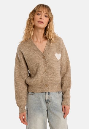 Donna che indossa un cardigan beige con bottoni e un cuore bianco sul petto e jeans azzurri, in piedi davanti a uno sfondo semplice.