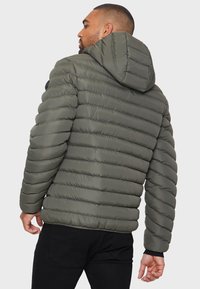 Puffjacka i mörk olivgrön färg, med quiltad textur, huva och ribbade ärmslut. Bakvyn visar framträdande horisontell söm.